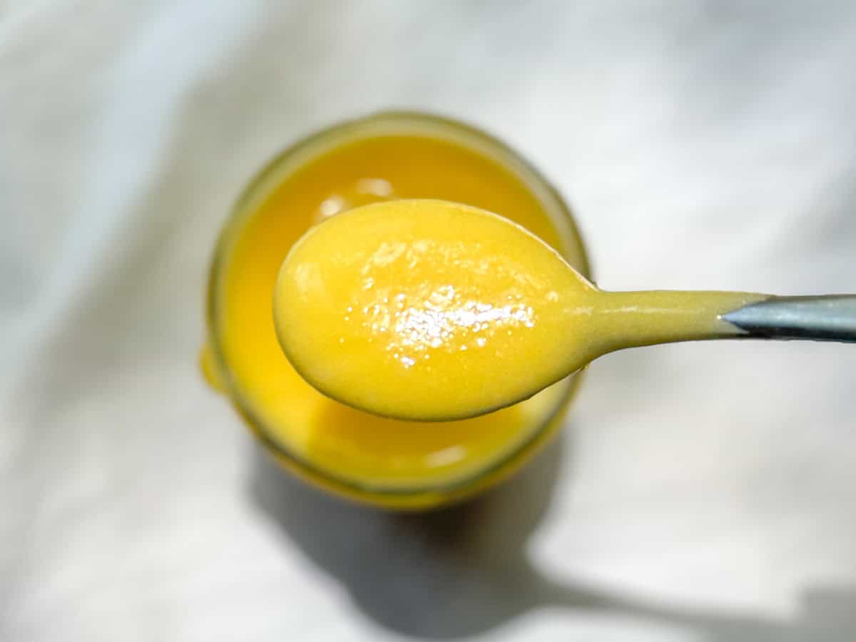 A spoon holding bright yellow easy lemon curd over a jar of more curd.