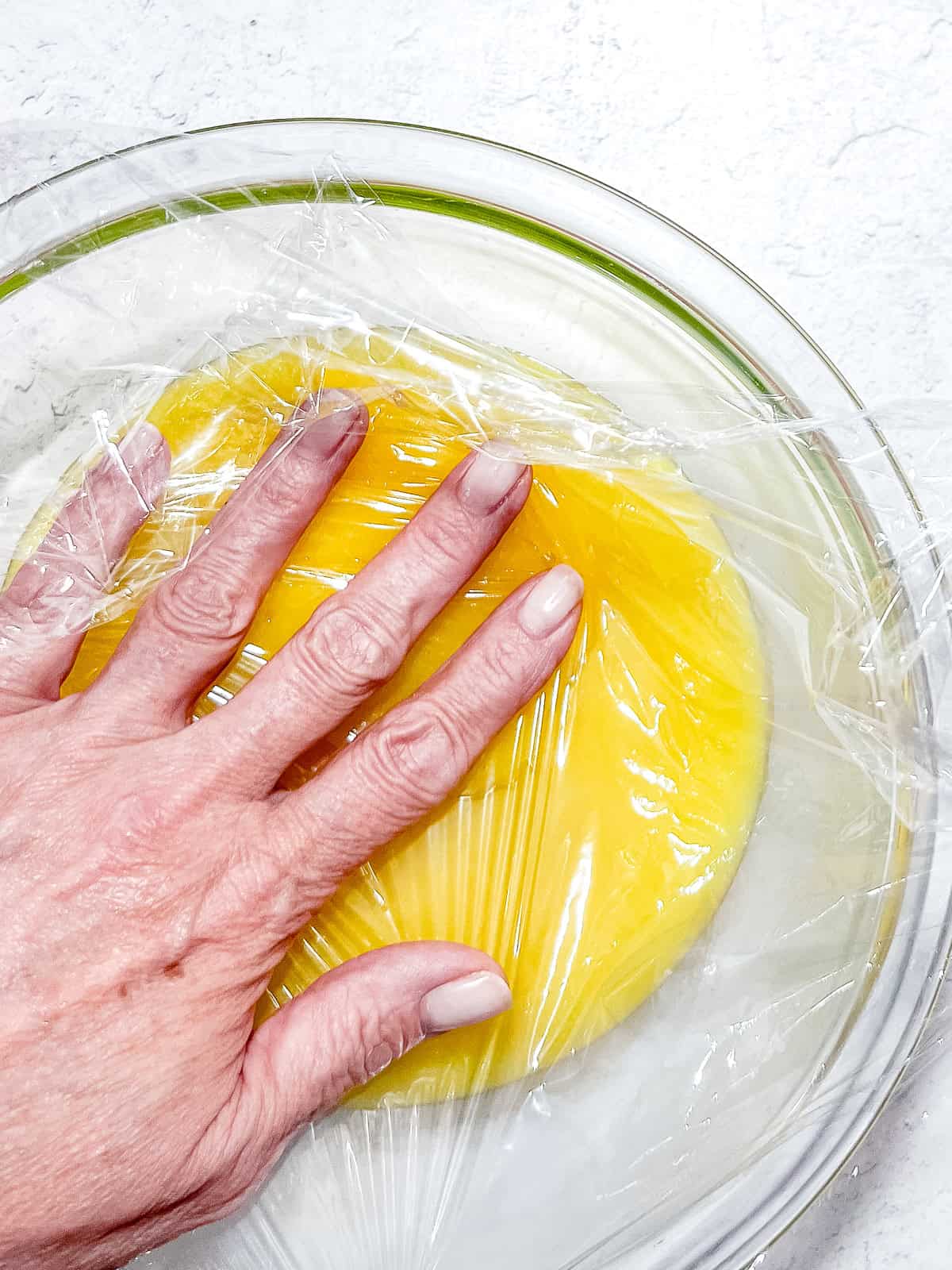 Pressing plastic cling film directly on the surface of curd in a glass bowl.