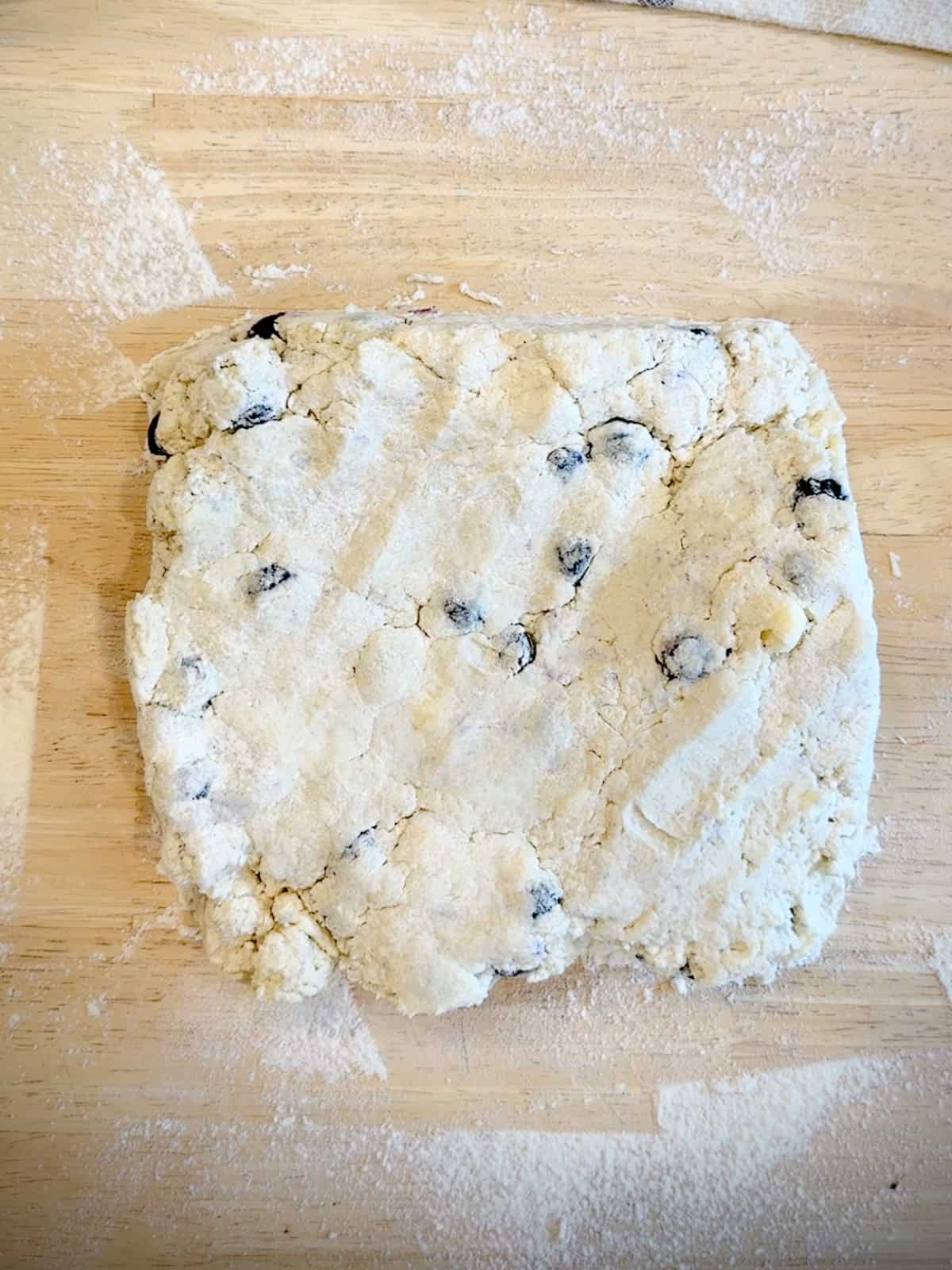 Shaping blueberry biscuit dough into a square on a wooden counter.