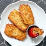 Three hash brown patties on a white plate with a cup of ketchup with a small piece of hash brown in it.