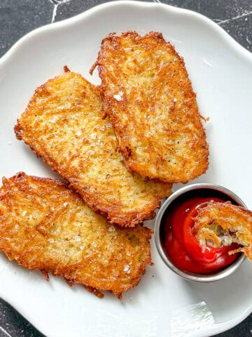 Three hash brown patties on a white plate with a cup of ketchup with a small piece of hash brown in it.
