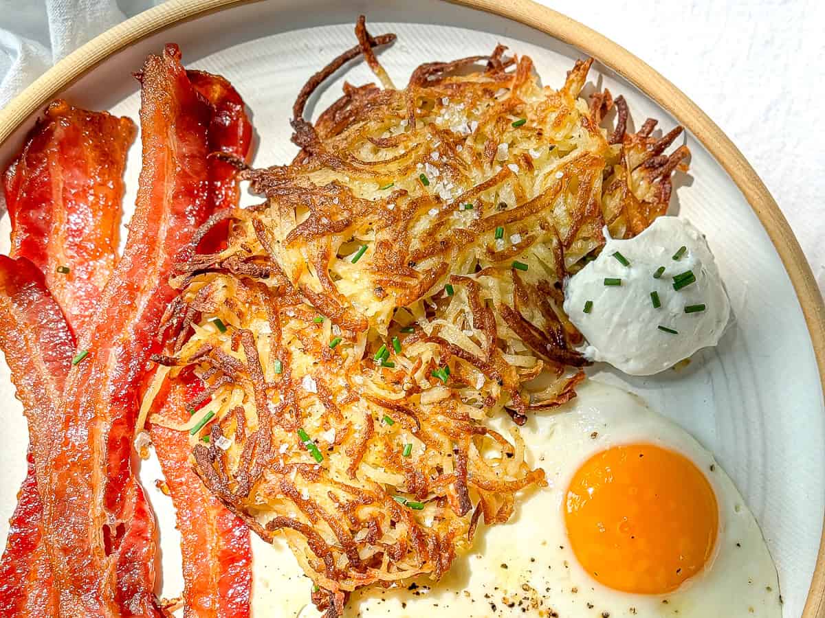 Hash brown patties with sour cream and chives on a white plate with bacon and eggs.