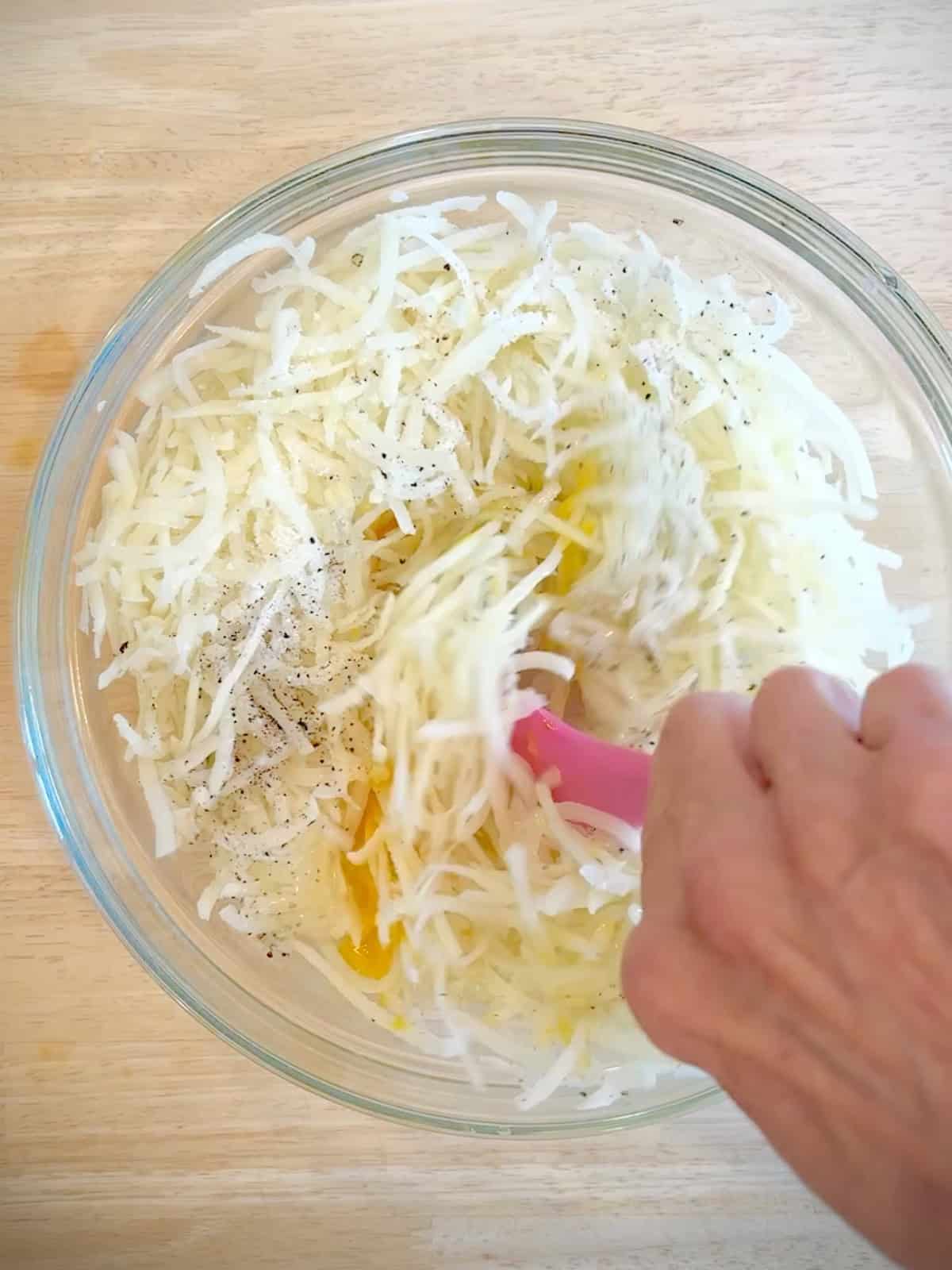 Mixing salt, pepper, egg, and flour into shredded potatoes in a glass bowl.