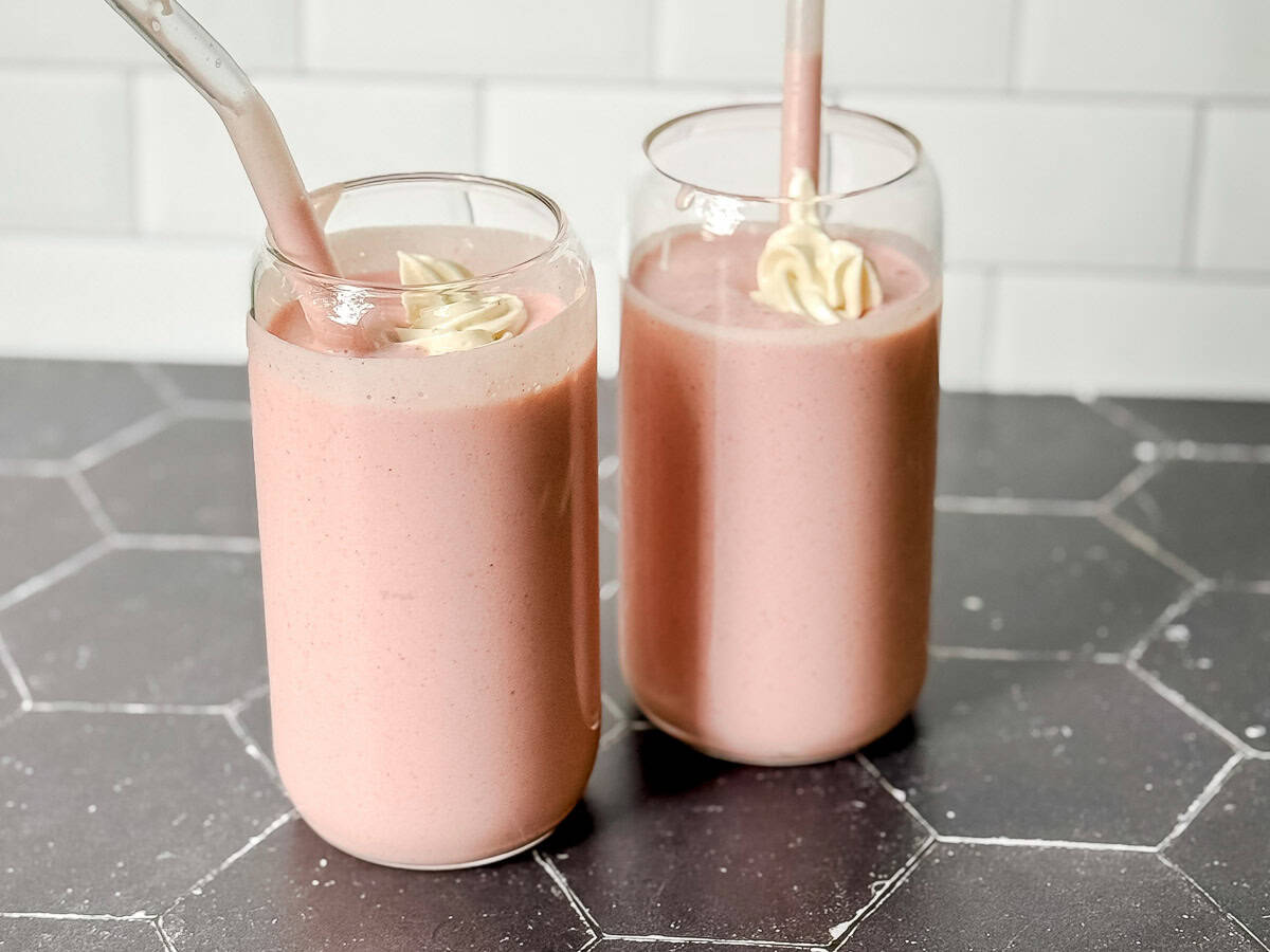 Two strawberry banana smoothies on a black background with a white wall behind it.
