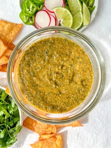 Roasted tomatillo salsa in a glass bowl with limes and cilantro in the background.