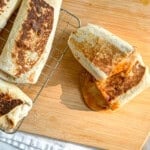 A bean and cheese burrito on a wooden cutting board with more burritos on a wire rack to the side.