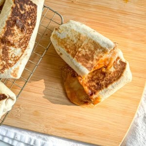 A bean and cheese burrito on a wooden cutting board with more burritos on a wire rack to the side.