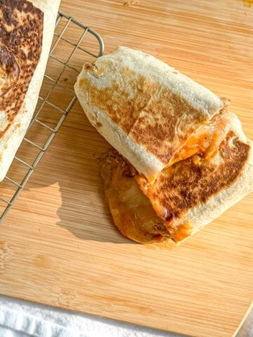 A bean and cheese burrito on a wooden cutting board with more burritos on a wire rack to the side.