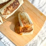 A bean and cheese burrito on a wooden cutting board with more burritos on a wire rack to the side.