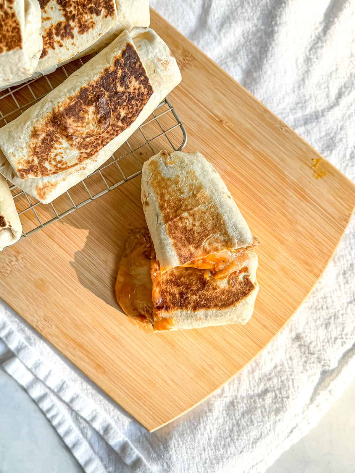 A bean and cheese burrito on a wooden cutting board with more burritos on a wire rack to the side.