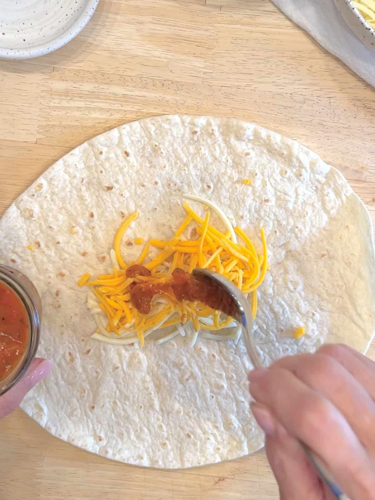 Placing cheese and ranchero sauce in a line across the center of a tortilla to make bean and cheese burritos.