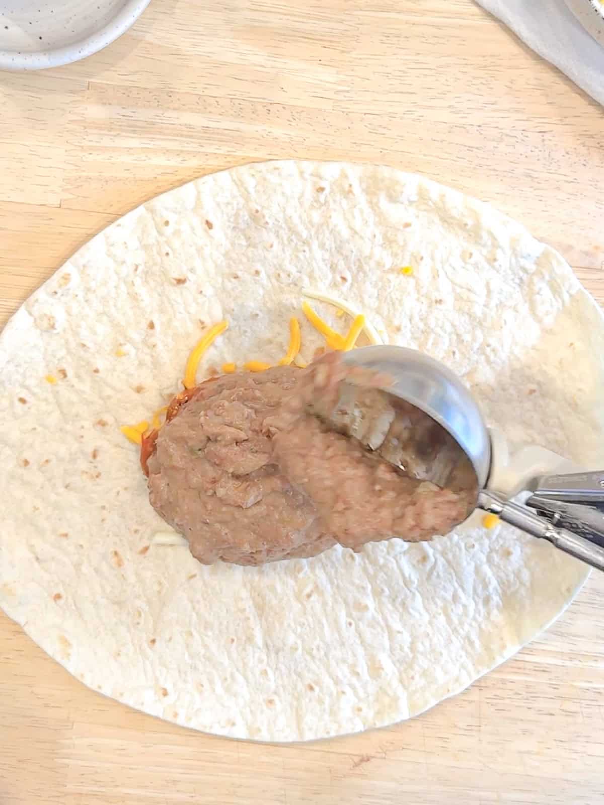 Arranging the refried beans on top of the cheese and sauce in a line across the center of a tortilla.