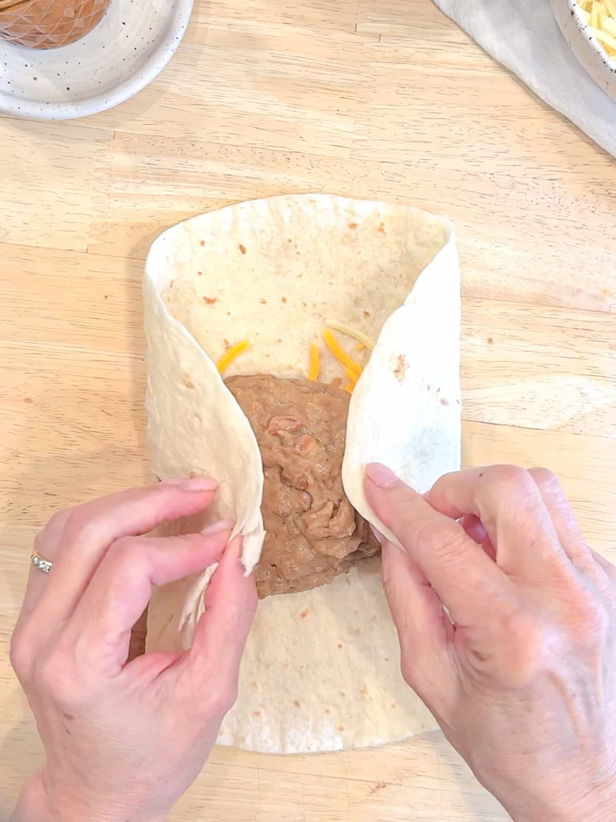 Folding the sides of a tortilla inwards, over the bean and cheese filling.