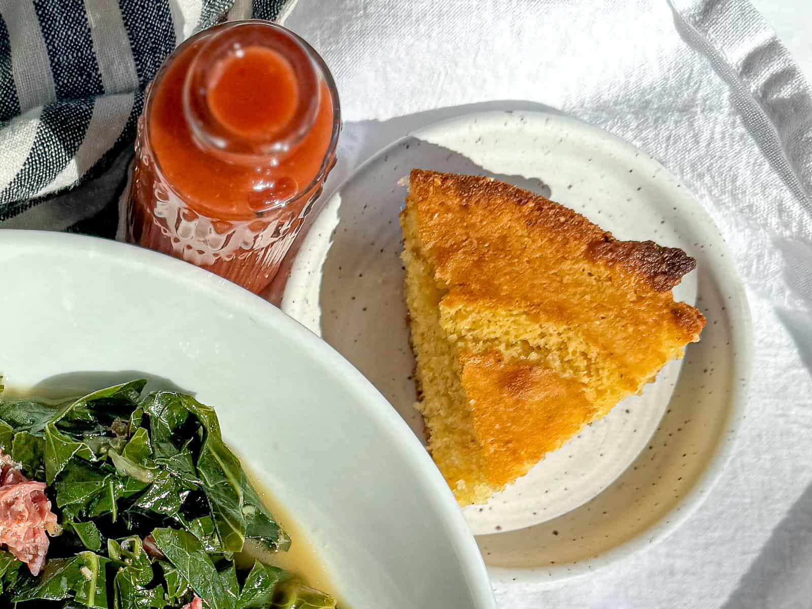 Collard greens in a white bowl with a dish of cornbread and a bottle of hot sauce nearby.