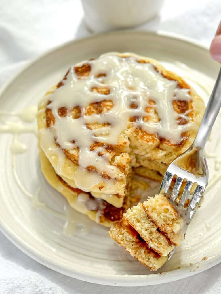 Using a fork to take a bite of a stack of cinnamon roll pancakes with cream cheese glaze.