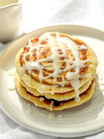 A stack of cinnamon roll pancakes with cream cheese glaze on a white plate.