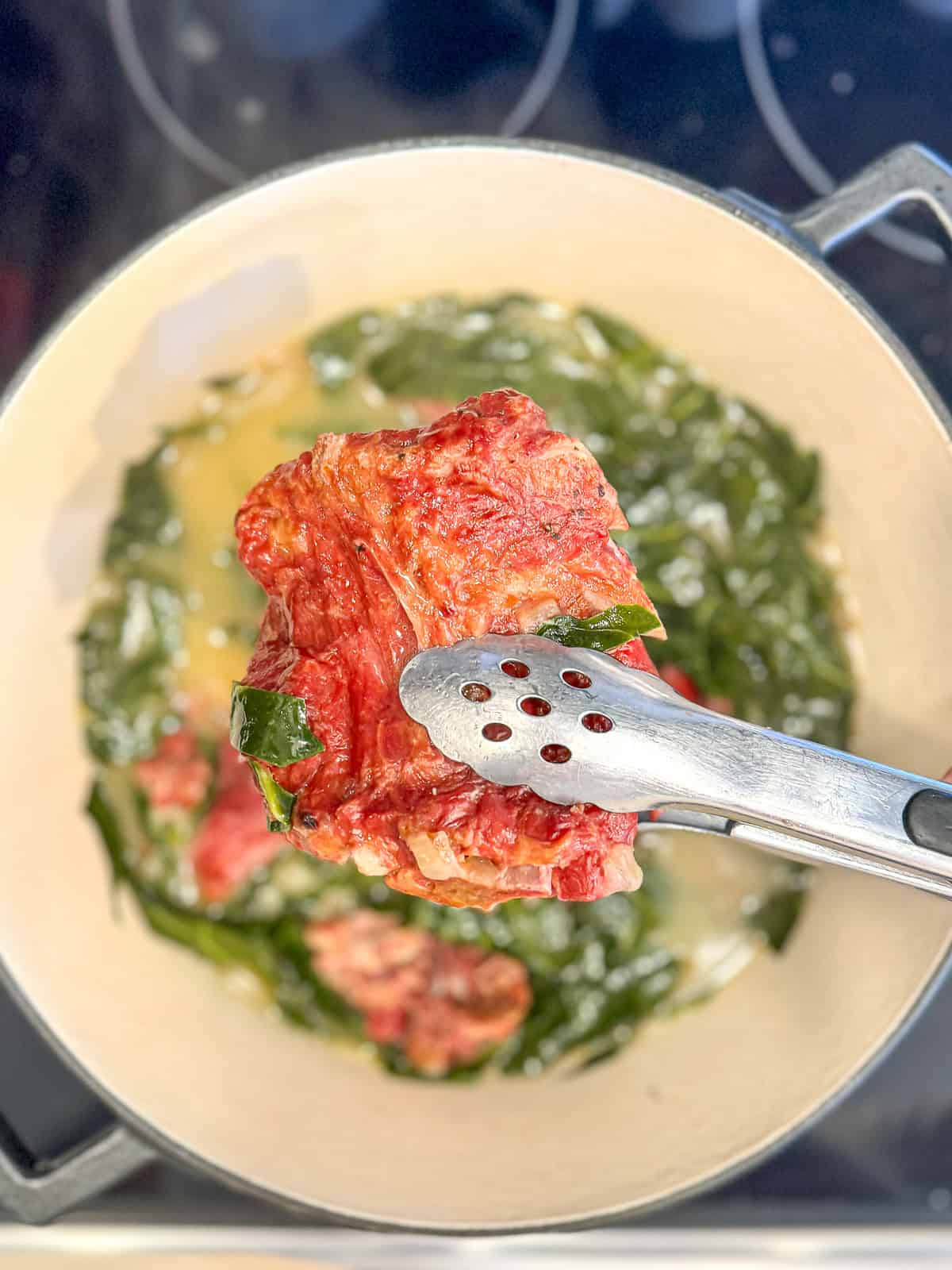 Using tongs to pull smoked pork out of a pot of collard greens for shredding.