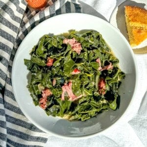 A white bowl filled with collard greens with smoked pork against a blue striped napkin with cornbread and hot sauce on the table.