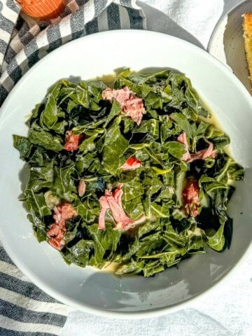 A white bowl filled with collard greens with smoked pork against a blue striped napkin with cornbread and hot sauce on the table.