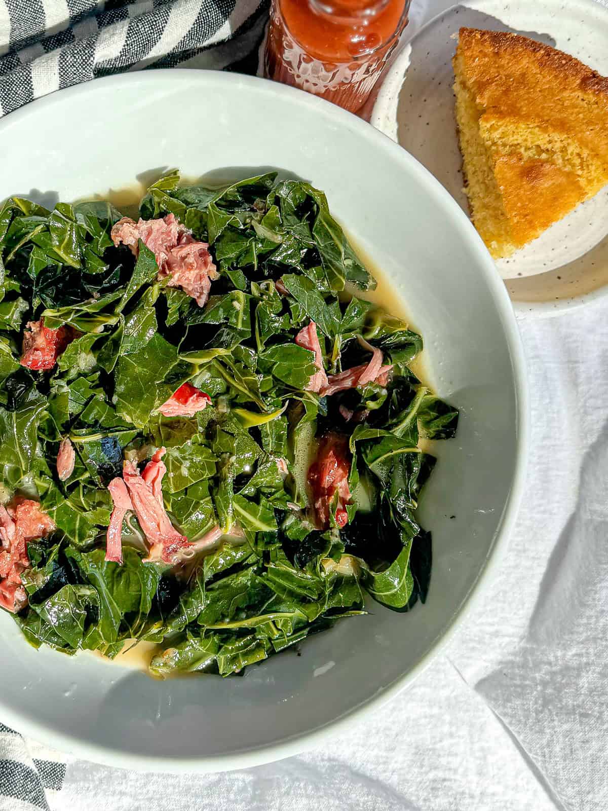 A white bowl filled with collard greens with smoked pork against a blue striped napkin with cornbread and hot sauce on the table.