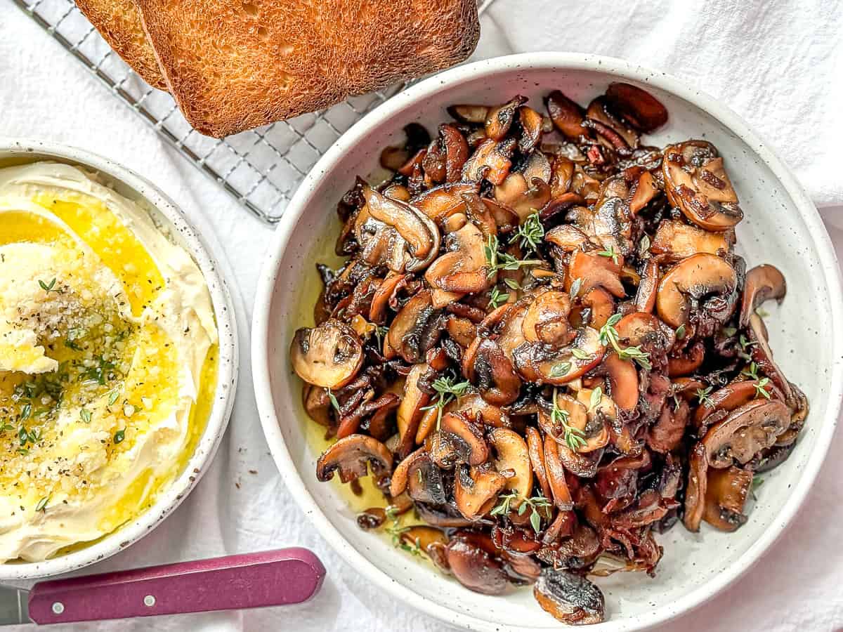 A bowl full of mushrooms, toasted, brioche bread, and cheesy mascarpone with olive oil and herbs on a white tablecloth.