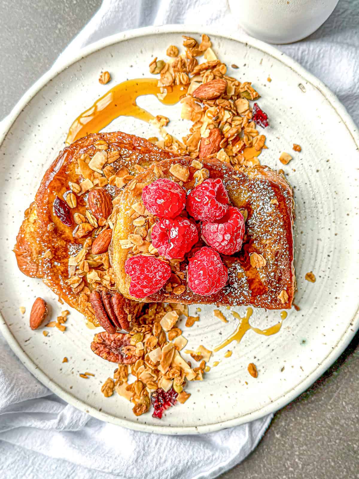 French toast with granola fresh fruit and maple syrup on a white plate.