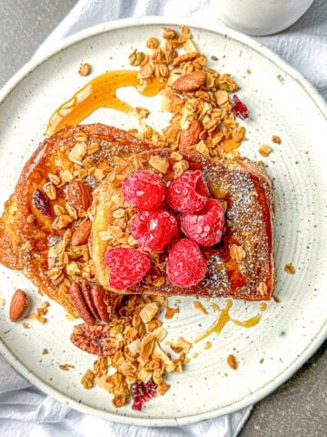 French toast on a white plate sprinkled with granola and fresh raspberries.