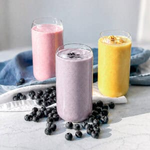 Three fruit smoothies on a marble counter with blueberries scattered on top of the counter.