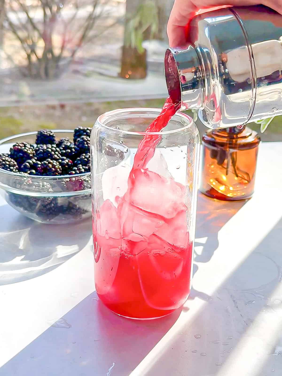 Pouring a shaken blackberry sage refresher from a cocktail shaker into a glass of ice.