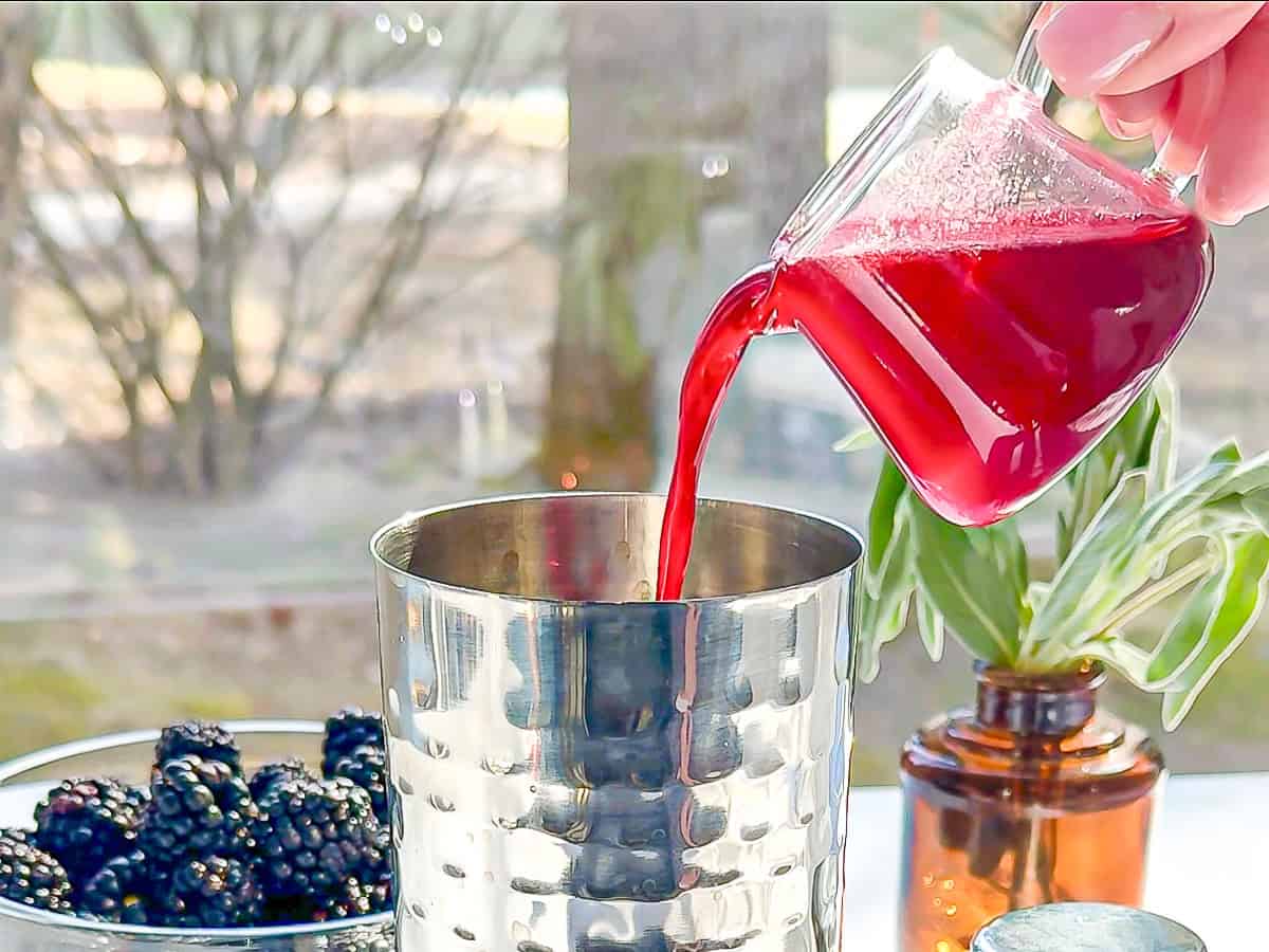 Pouring blackberry sage simple syrup into a cocktail shaker.