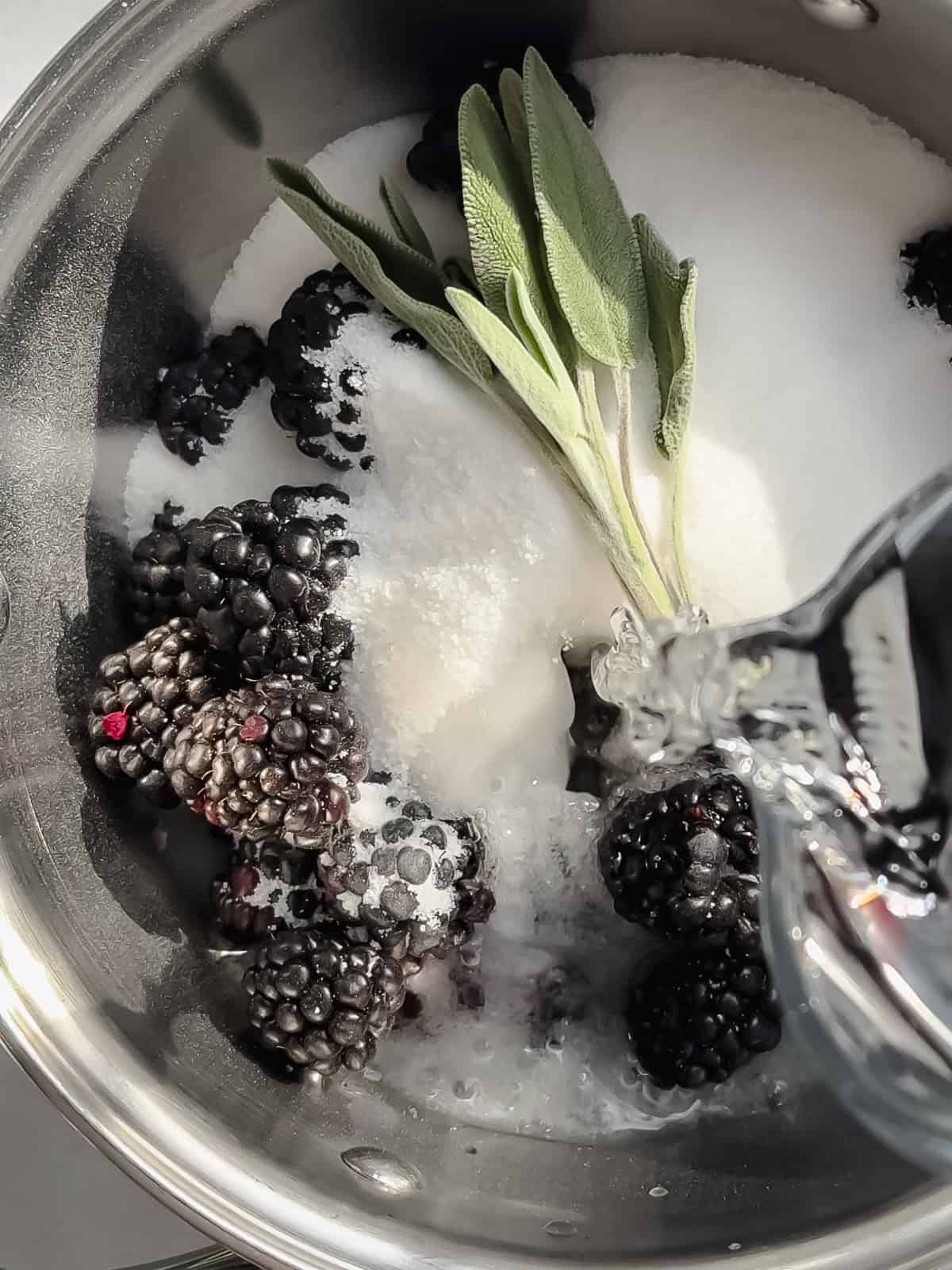 Pouring water into a saucepan containing blackberries, sugar, and sage leaves to make blackberry sage simple syrup.