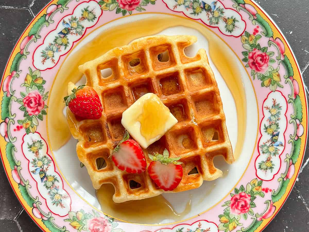 A waffle with butter syrup and strawberries on a floral plate.