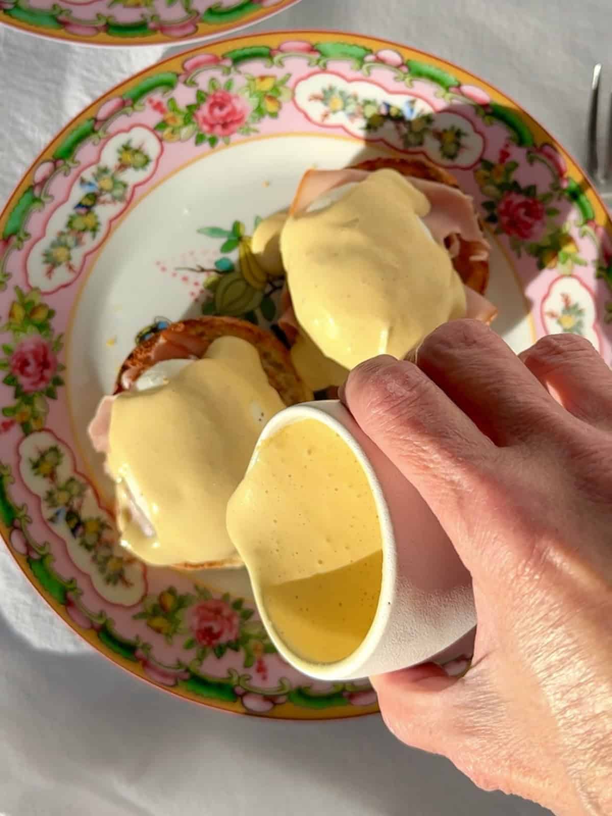 Pouring hollandaise sauce over poached eggs, ham, and an English muffin.