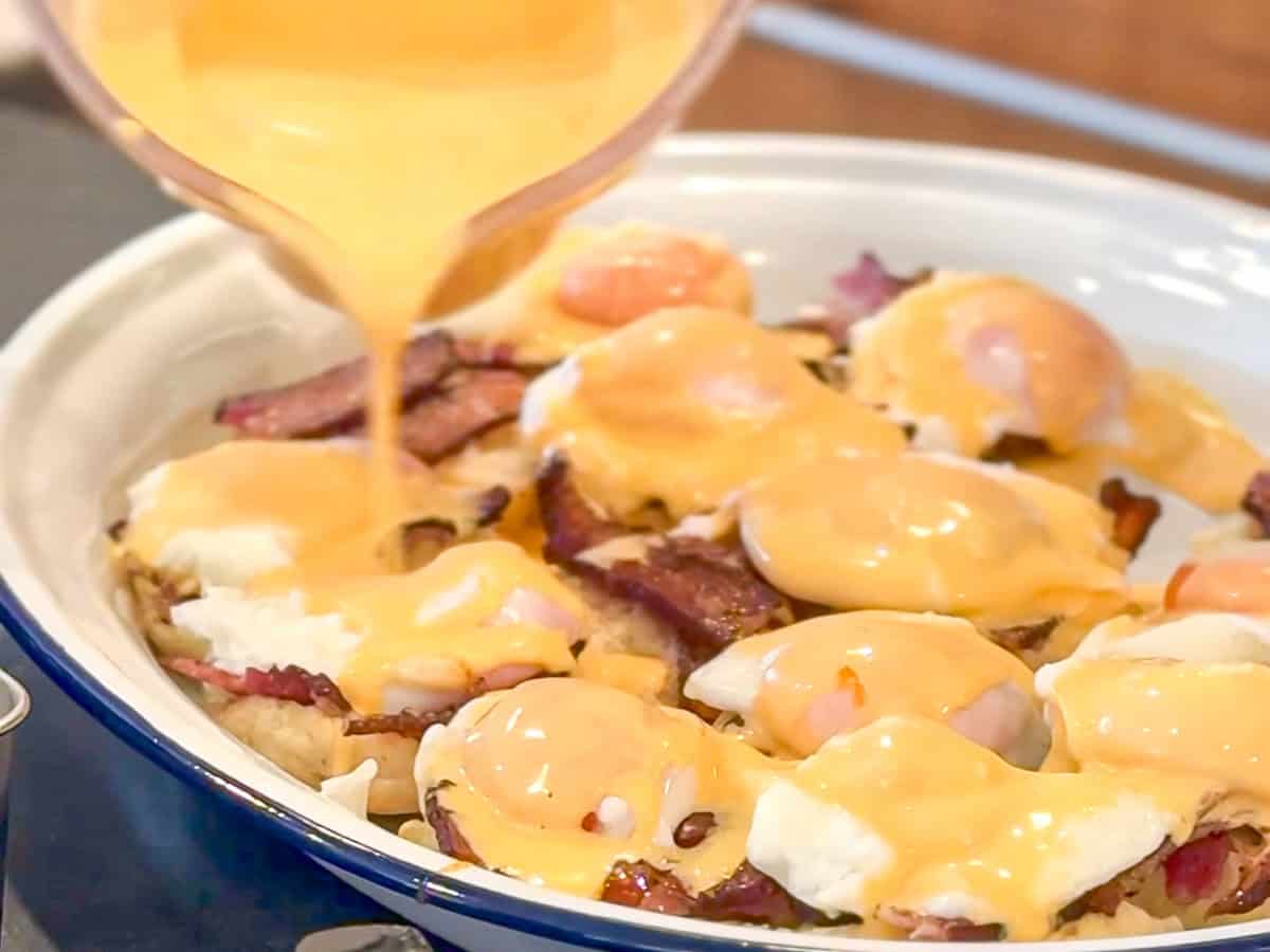 Pouring hollandaise sauce over a tray containing a lerge number of eggs benedict.
