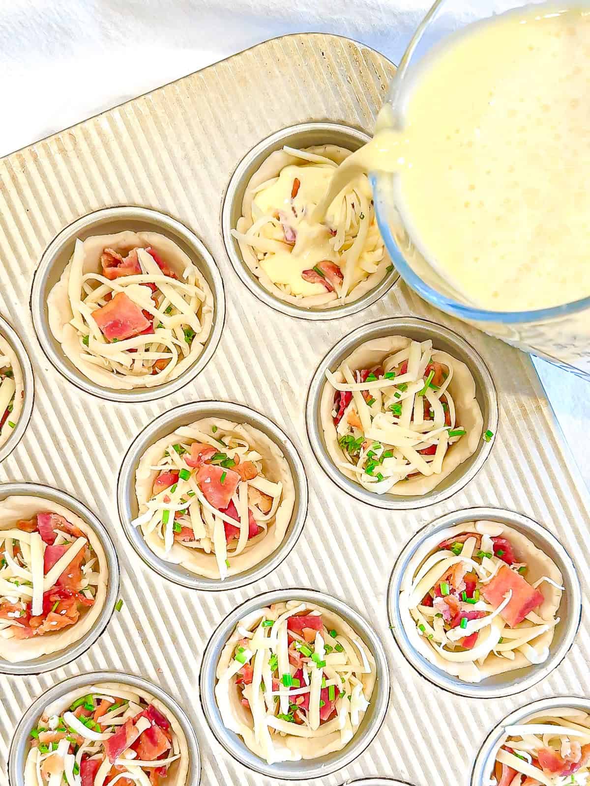 Pouring egg mixture over ingredients in a pie shell in a muffin pan.