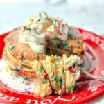 A fork holding birthday cake pancakes, with the full stack in the background with whipped cream and sprinkles on top.