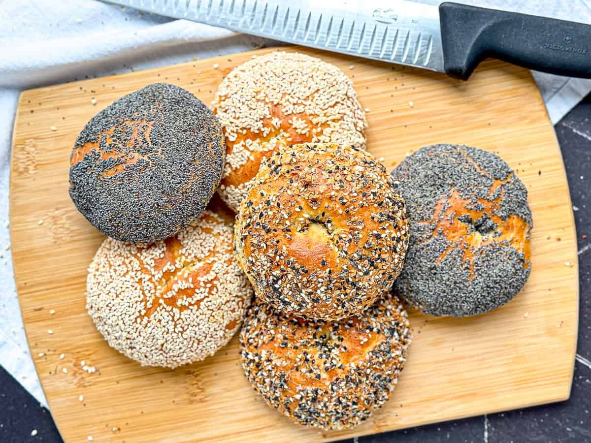 A stack of cottage cheese bagels with various toppings; sesame seeds, poppy seeds, and everything bagel seasoning, on a wooden cutting board.