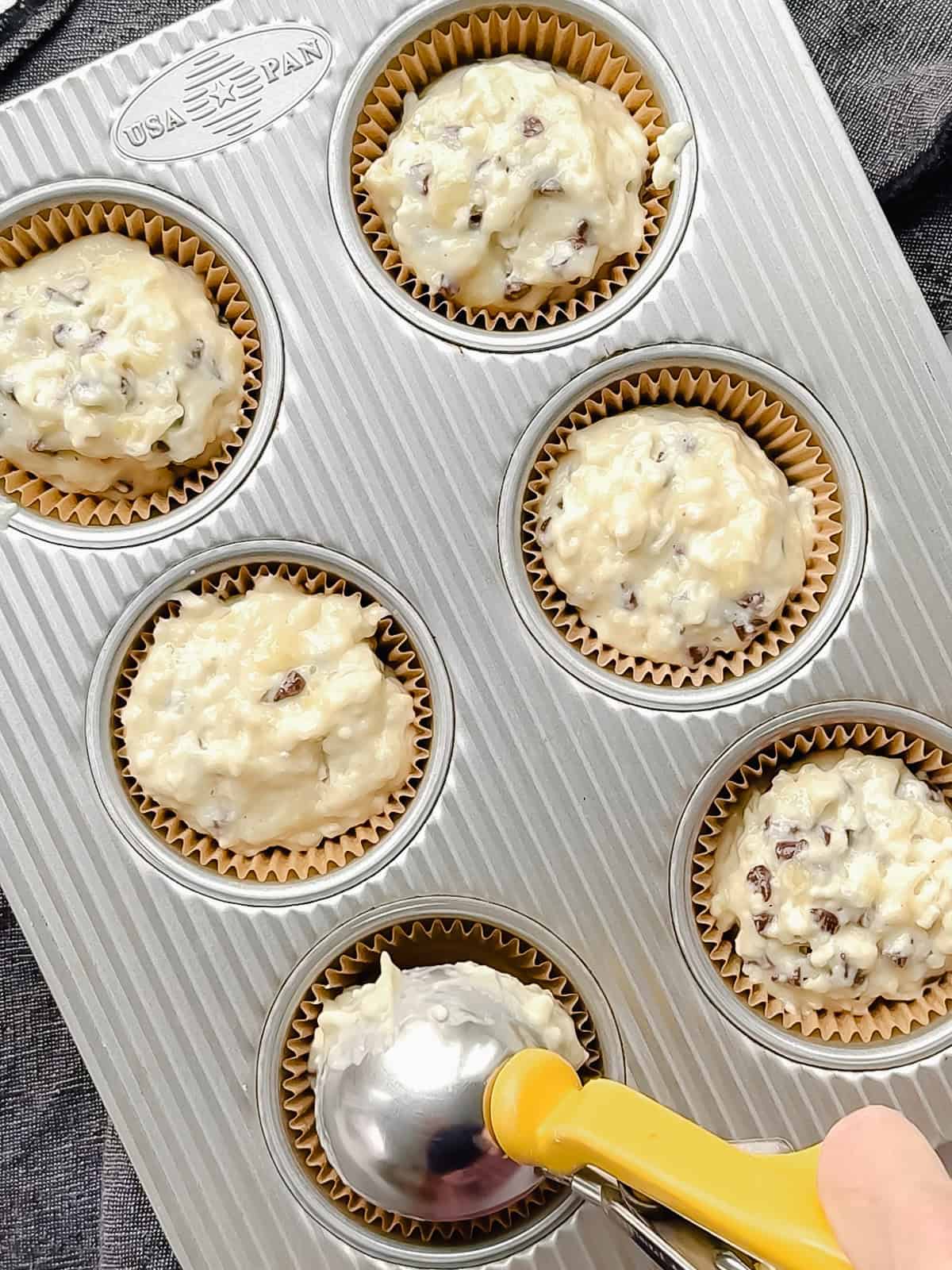Using an ice cream scoop to portion muffin batter into a paper lined muffin pan.