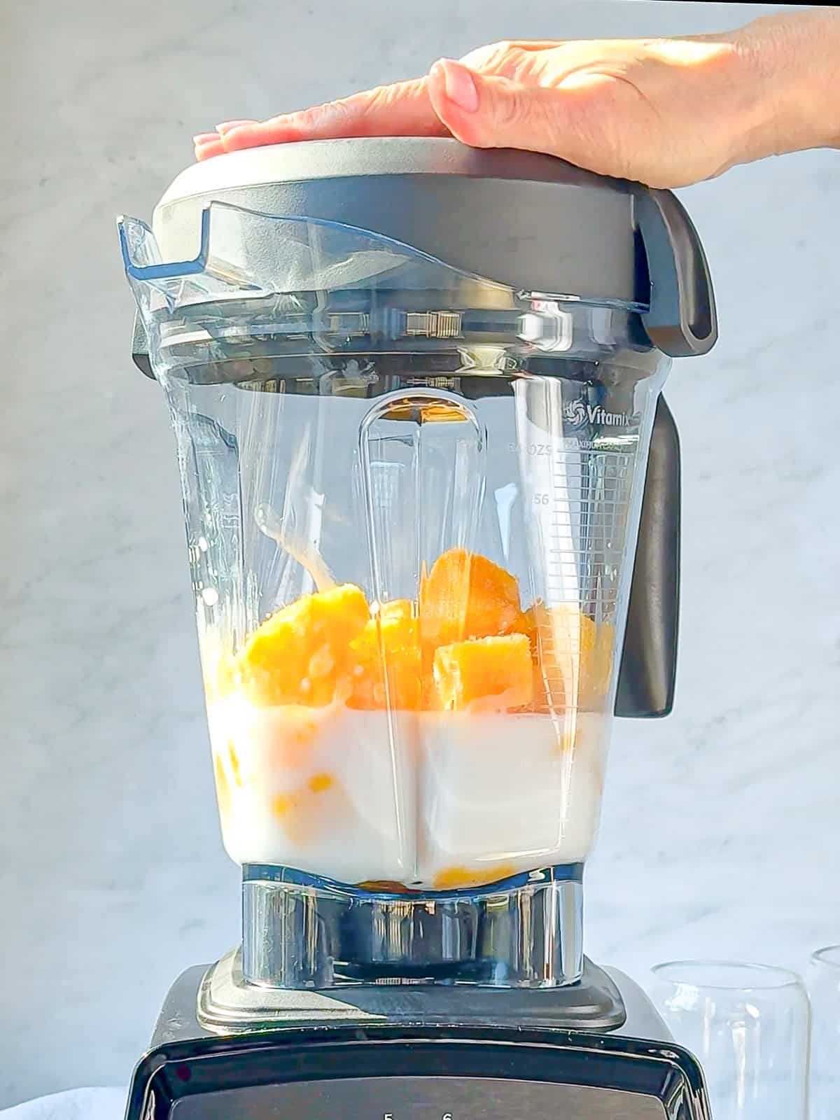 A hand holds the lid of a blender filled with chunks of mango and a creamy liquid, set against a light background. Its poised to blend everything into a refreshing mango yogurt smoothie.