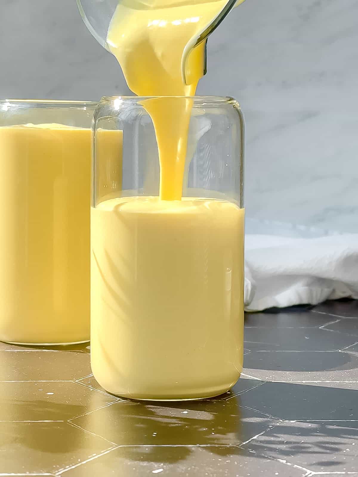 A glass is being filled with a creamy, smooth mango yogurt smoothie from a pitcher. Another filled glass sits in the background, set against a light backdrop with natural lighting.