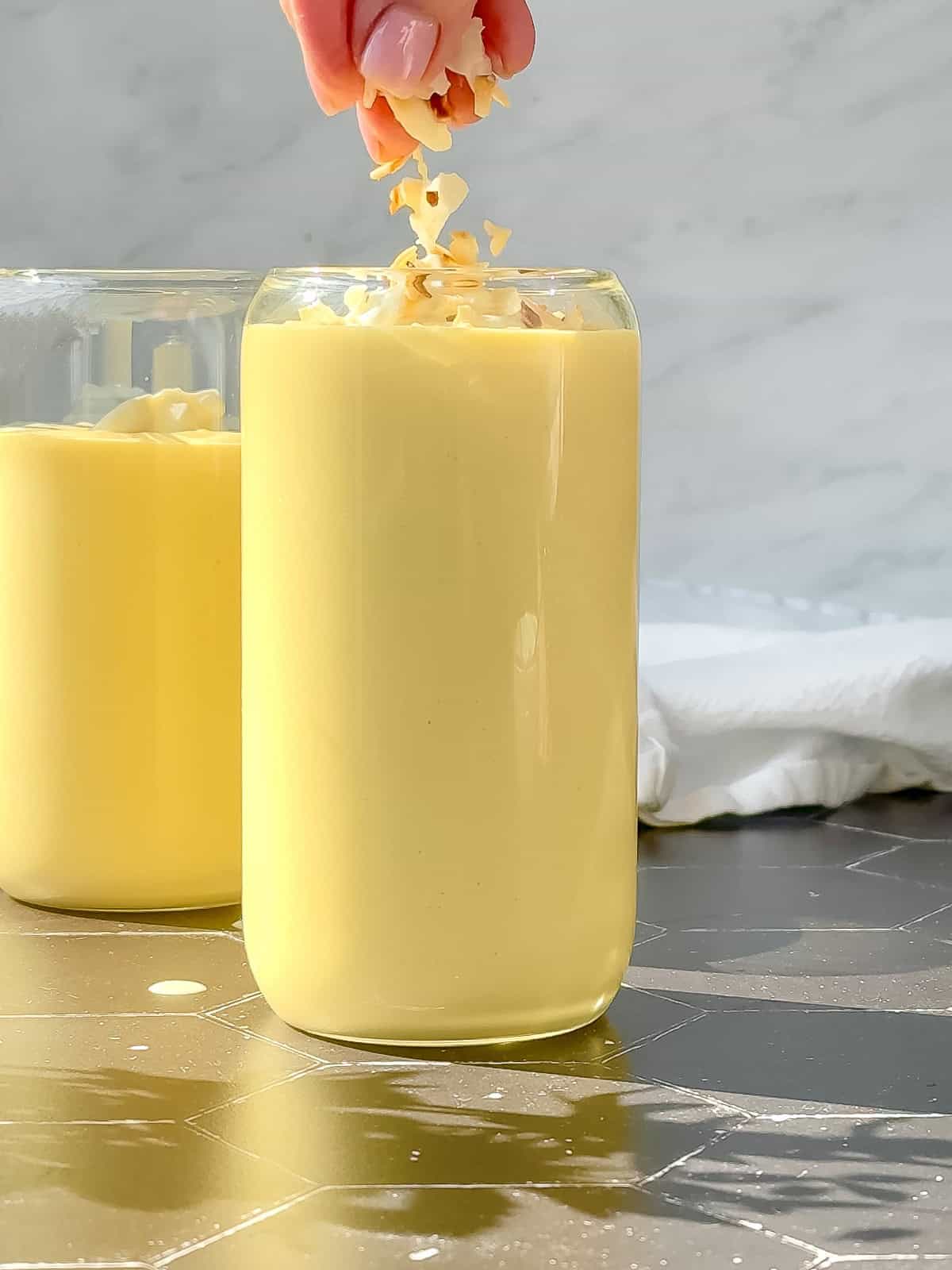 A hand sprinkles toasted coconut over a tall glass filled with a creamy mango yogurt smoothie. Another glass of the luscious drink is slightly blurred in the background, set on a dark, speckled surface against a light marble backdrop.