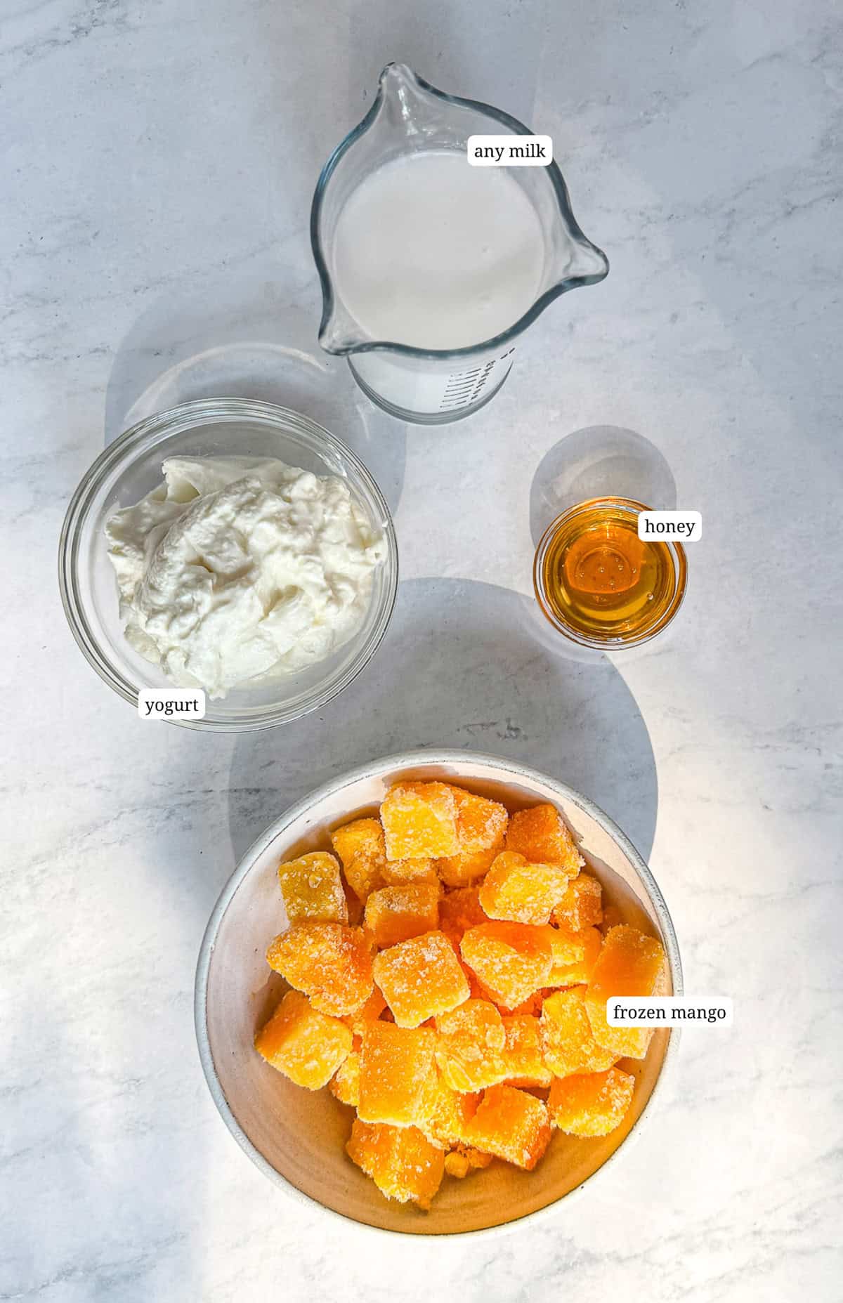 A top-down view of smoothie ingredients on a white marble surface: a bowl of frozen mango chunks, a small glass of honey, a clear jug of milk, and a bowl of yogurt.