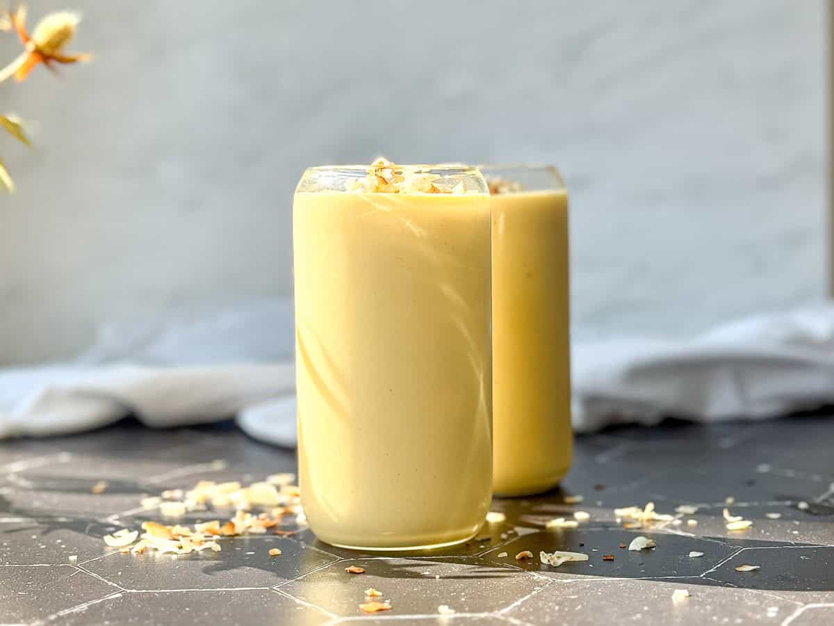 Two glasses of creamy yellow mango yogurt smoothie topped with toasted coconut flakes rest on a hexagonal tiled surface.