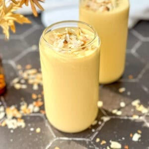 Two glasses of creamy yellow mango yogurt smoothie topped with toasted coconut flakes rest on a hexagonal tiled surface.