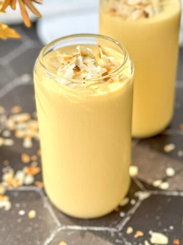 Two glasses of creamy yellow mango yogurt smoothie topped with toasted coconut flakes rest on a hexagonal tiled surface.