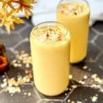 Two glasses of creamy yellow mango yogurt smoothie topped with toasted coconut flakes rest on a hexagonal tiled surface.