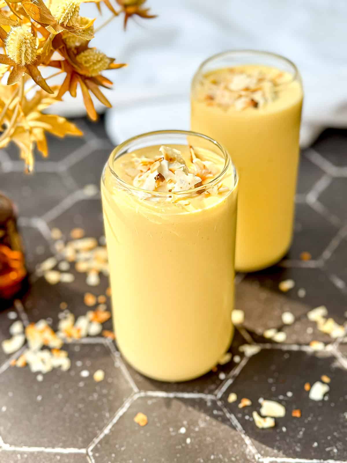 Two glasses of creamy yellow mango yogurt smoothie topped with toasted coconut flakes rest on a hexagonal tiled surface.