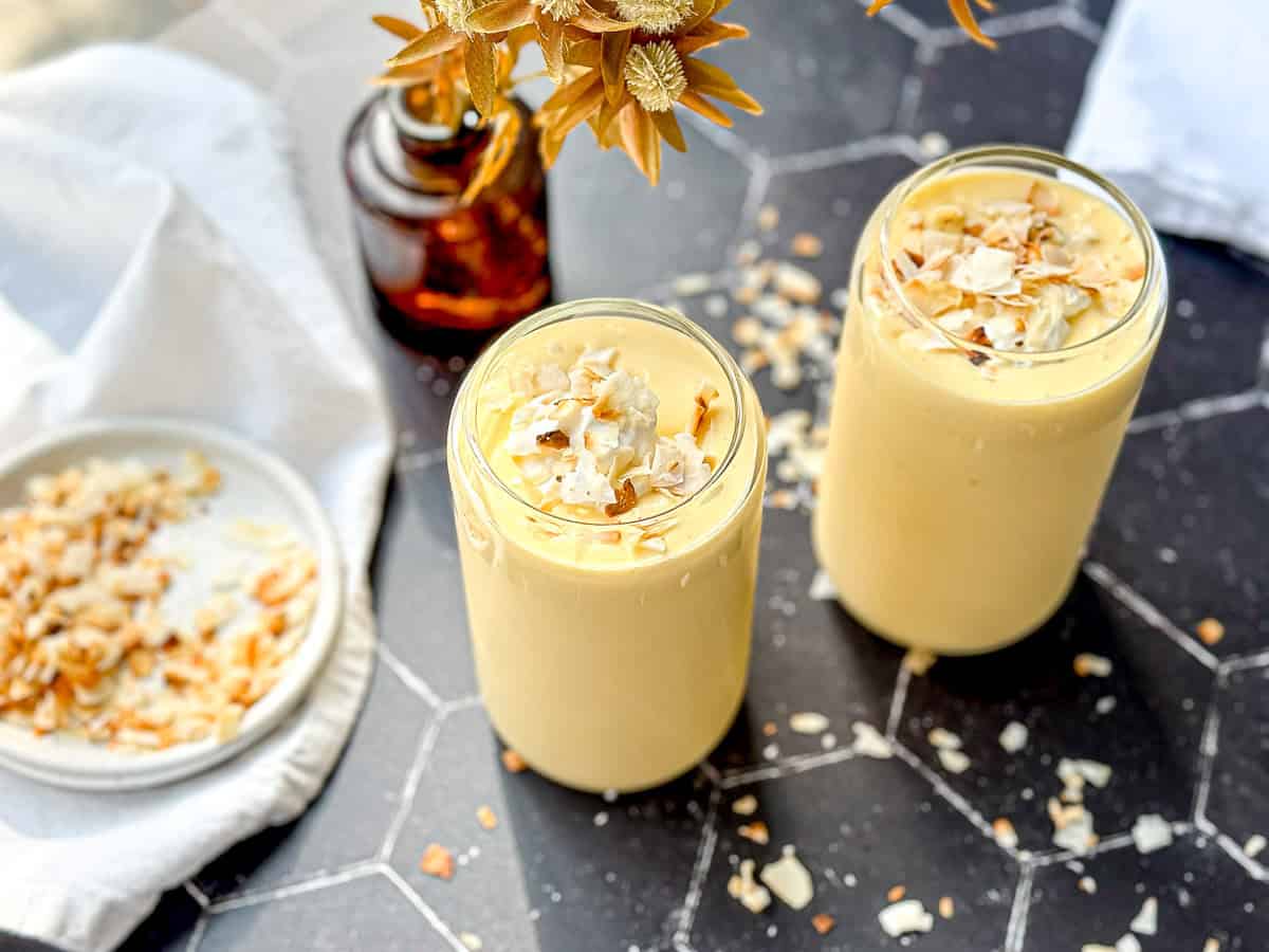 Two glasses of creamy yellow mango yogurt smoothies topped with coconut flakes sit on a black hexagonal-tile table.