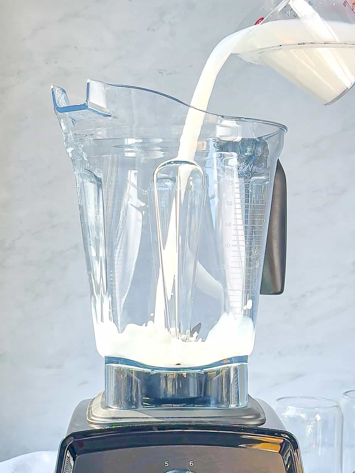 A person pours milk from a measuring cup into a transparent blender container, which is partially filled with milk to prepare a mango yogurt smoothie.