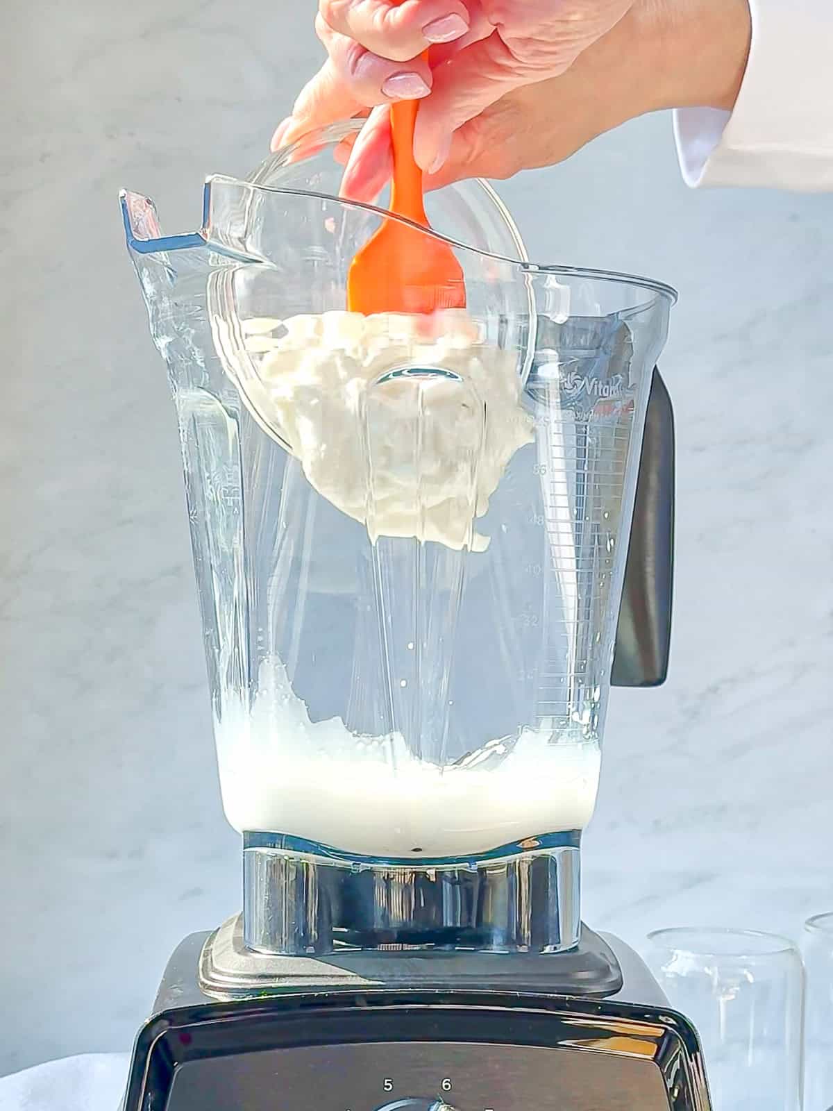 A hand is holding a spatula, using it to add creamy yogurt from a glass bowl into an electric blender. The blender base is visible, along with some white liquid already inside the jar—perhaps the beginnings of a mango yogurt smoothie.